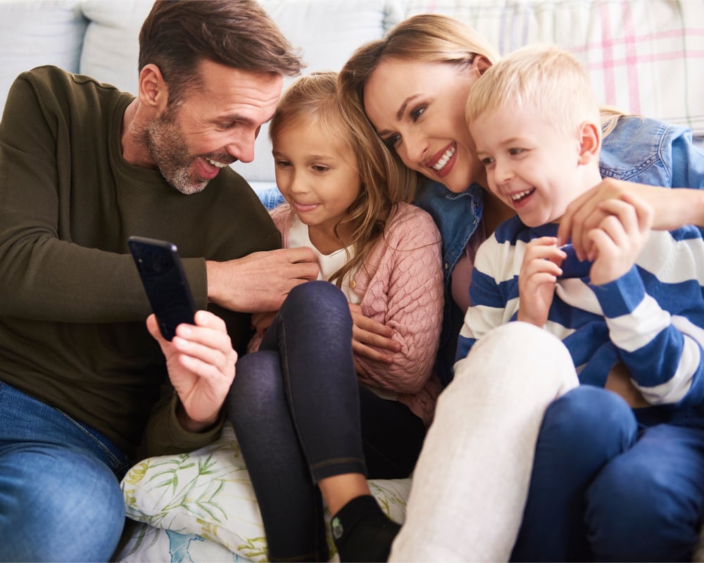 Une famille de quatre personnes est assise ensemble sur un canapé en regardant un téléphone.